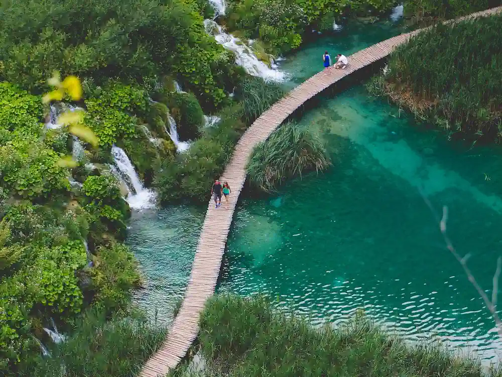這張圖片展現了克羅地亞深度遊的必訪景點 十六湖國家公園(Plitvice Lakes National Park)的壯麗景色。從高處俯瞰,一條木製棧道蜿蜒穿過清澈見底的翠綠色湖泊,周圍是茂密的森林和層疊的小型瀑布。幾名遊客正在棧道上漫步,近距離體驗這片原始的自然美景。此圖片適用於介紹克羅地亞自由行、巴爾幹半島旅遊、國家公園攻略或深度遊行程的文章。
