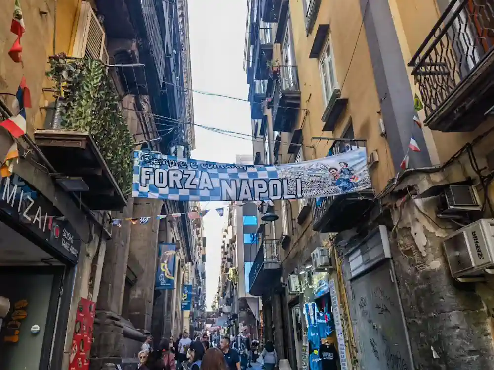 意大利拿坡里一條充滿遊客的歷史舊城區街道，街道上方掛著一幅藍白色的「Forza Napoli」足球應援橫額。