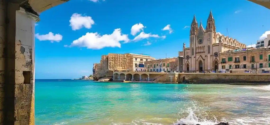 從拱門眺望馬爾他聖朱利安的巴魯塔灣，海水清澈碧藍，背景是宏偉的加爾默羅山聖母教堂。