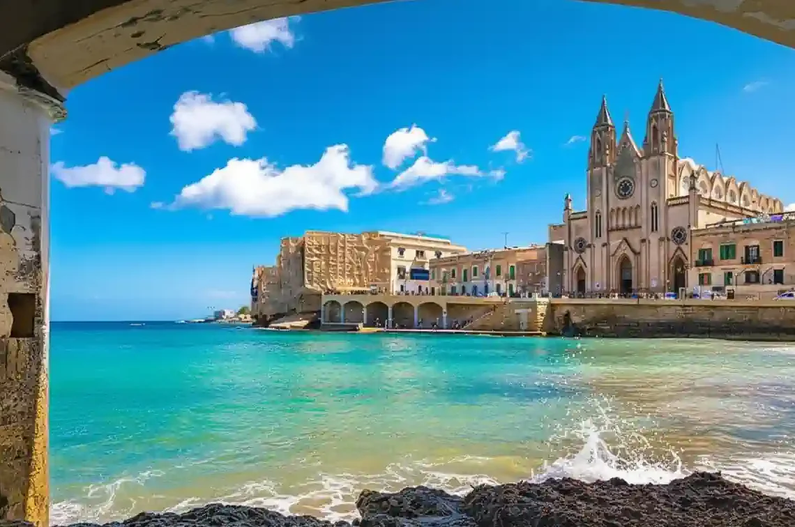 從拱門眺望馬爾他聖朱利安的巴魯塔灣，海水清澈碧藍，背景是宏偉的加爾默羅山聖母教堂。